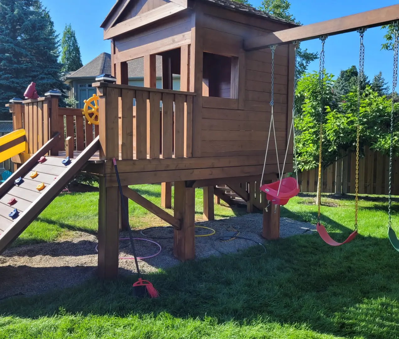 A stained cedar deck structure in Toronto: horizontal surfaces take direct overhead UV, standing water, and foot traffic that vertical fences never see Stained cedar deck and play structure in a Toronto backyard under full summer sun